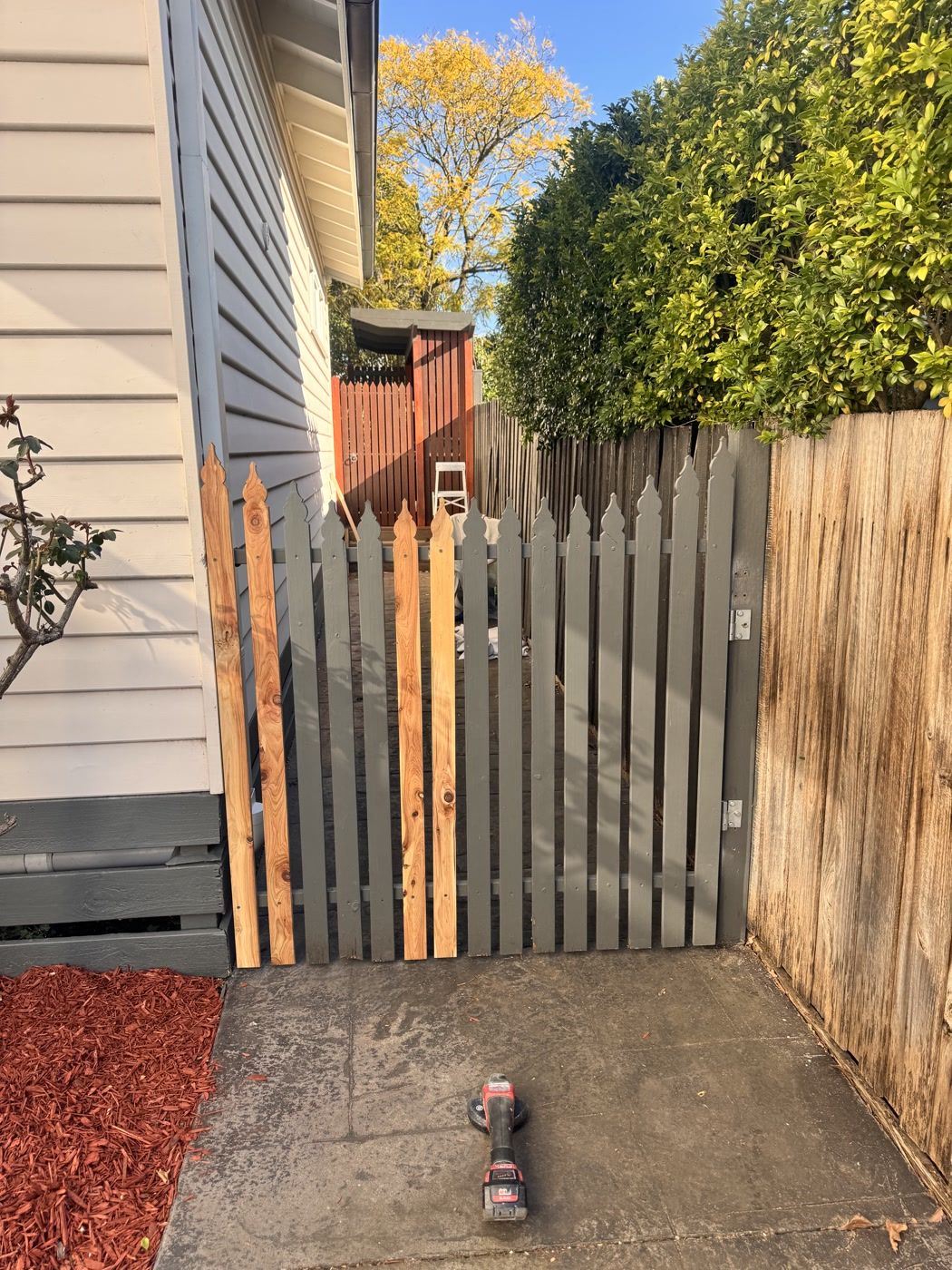 Dark grey picket gate with several new unpainted timber pickets installed